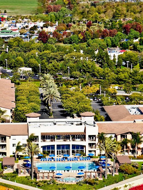 Aerial view of a resort with a large hotel, pool, palm trees, landscaped grounds, and colorful flower beds in the foreground and surrounding greenery.