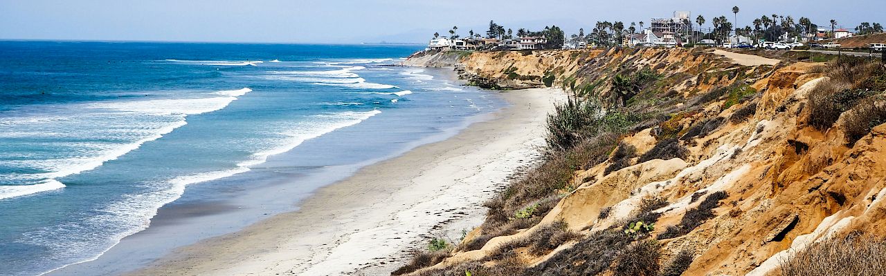 Crescent beach with gentle waves, a sandy shore, and rugged cliffs; distant houses line the horizon under a clear, blue sky.