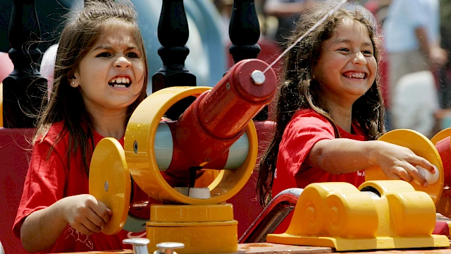 Two smiling girls in red shirts play with bright yellow water cannons and a mechanical toy on a sunny outdoor ride.