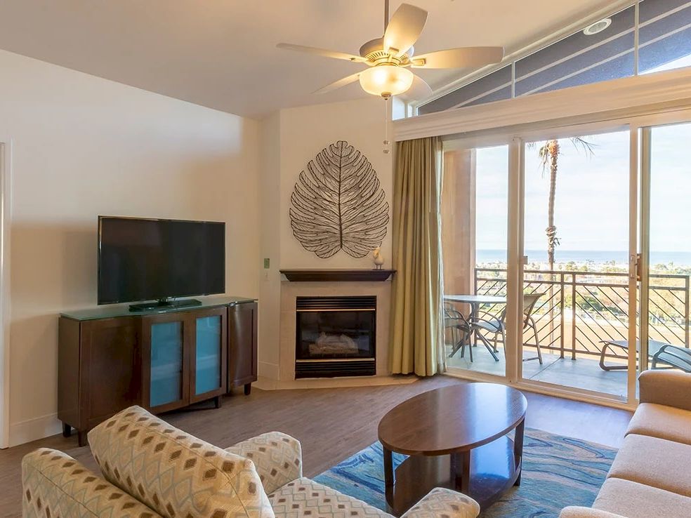 A cozy hotel suite with a living area, fireplace, TV on a cabinet, and a balcony with a view; separate bedroom visible in the doorway.