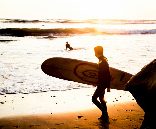 Two surfers walk along a sunny beach at sunset, carrying a board toward the water.