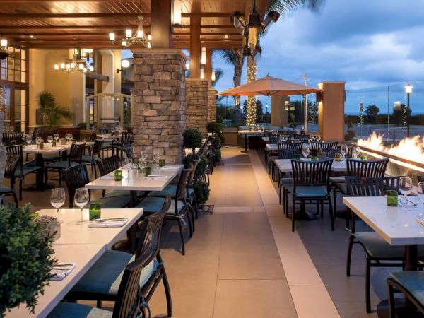 A cozy outdoor patio dining area with set tables, warm string lights, and a glowing fire pit on a nighttime terrace.